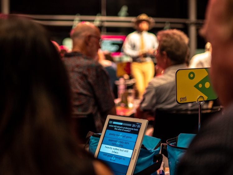 Image of A tablet displays trivia questions on a table among seated people at an event. In the background, a host stands and speaks, slightly blurred, whilst attendees listen and watch.