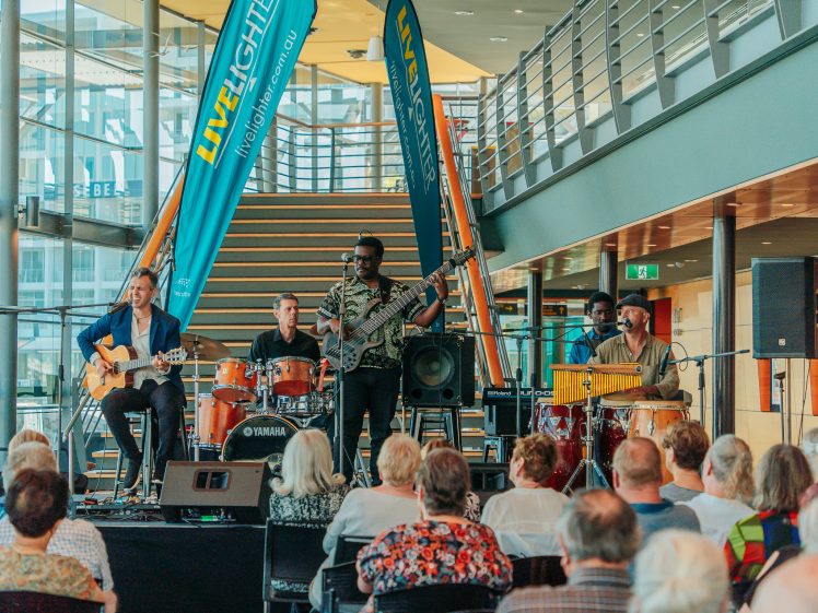 Image of A five-member band performs on stage with guitars, drums, and percussion instruments before an audience in a modern indoor venue with stairs and large windows in the background.