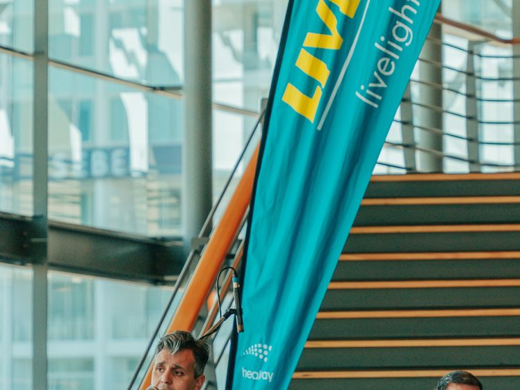 Image of A man seated and playing an acoustic guitar sings into a microphone next to a drummer. They perform indoors near a staircase beside a large blue “LiveLighter” banner.