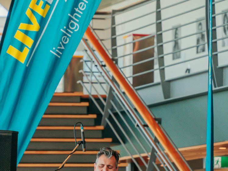 Image of A man in a blue blazer plays an acoustic guitar and sings into a microphone onstage, with a drummer behind him. A blue LiveLighter banner and a modern stairway are visible in the background.