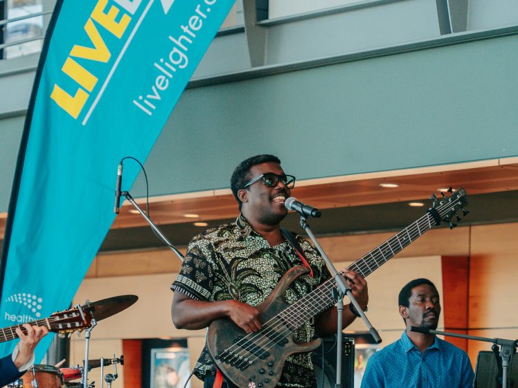 Image of A musician plays bass guitar and sings into a microphone on stage, accompanied by a keyboard player and drummer. They perform indoors near a large blue LiveLighter banner, with an audience watching.