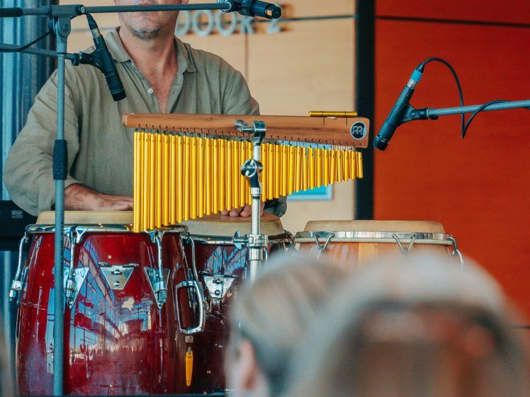 Image of A man wearing a flat cap plays conga drums and chimes onstage, with microphones set up in front of him. An audience watches, and a sign with 18–34 is visible in the background.