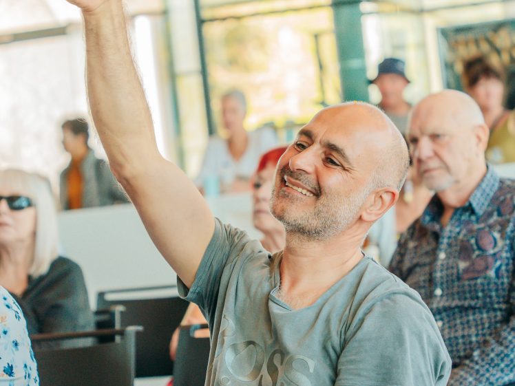 Image of A smiling man sitting in a chair raises his arm and points upwards, appearing engaged in a lively discussion or activity. Other seated people watch attentively in the background, in a bright indoor setting.