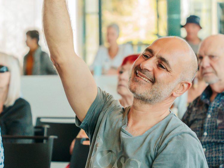 Image of A smiling man with a shaven head sits in a chair and raises his arm, pointing upwards, in a bright indoor setting with other seated people in the background.