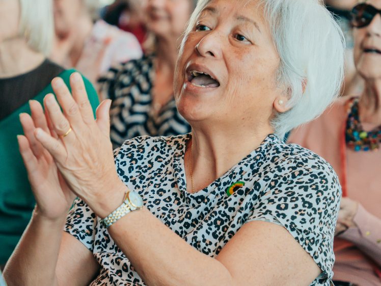 Image of An elderly woman with white hair and a leopard-print top claps her hands and smiles in a crowd of seated people at an indoor event.