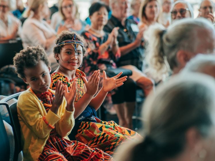 Image of Two young children in colourful traditional clothing sit and clap at an indoor event, surrounded by an audience of adults who are also clapping and watching attentively.