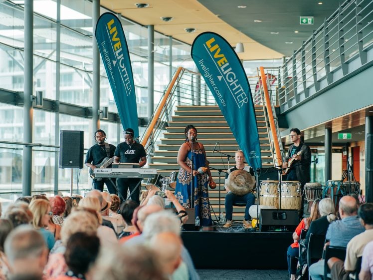 Image of A band performs live indoors in front of an audience. The group includes a singer, keyboard player, drummer, and percussionists. Two large LiveLighter banners stand behind them near a staircase.
