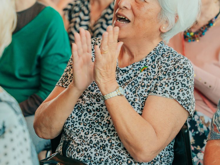 Image of An older woman with white hair claps enthusiastically whilst sitting in a crowd. She is smiling and wearing a leopard-print top, with other people seated and watching in the background.
