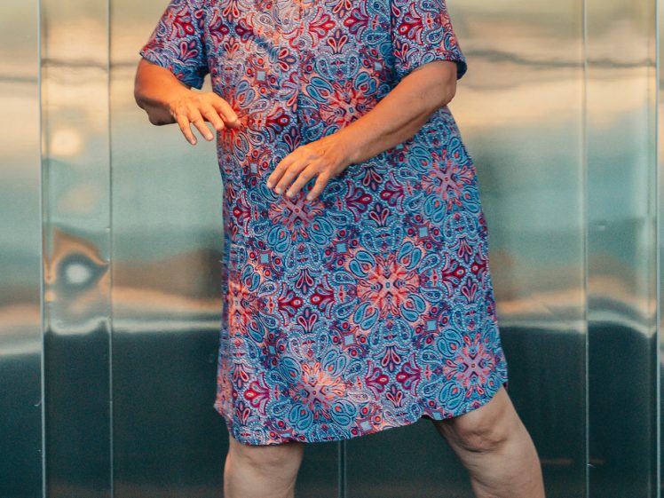 Image of A person with short hair, wearing glasses and a colourful patterned dress, stands smiling with arms playfully positioned, in front of a shiny metallic background.