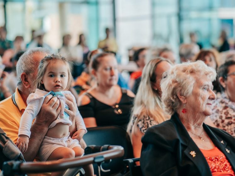 Image of A toddler sits on an older adult's lap in a crowded indoor setting, surrounded by people of various ages who are seated and attentively facing forwards. The scene appears to be at a public event or gathering.