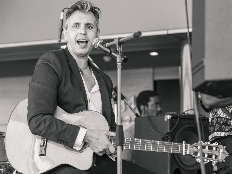Image of A man in a suit sits and plays an acoustic guitar while singing into a microphone on stage. Drums and another musician are visible in the background. The image is in black and white.