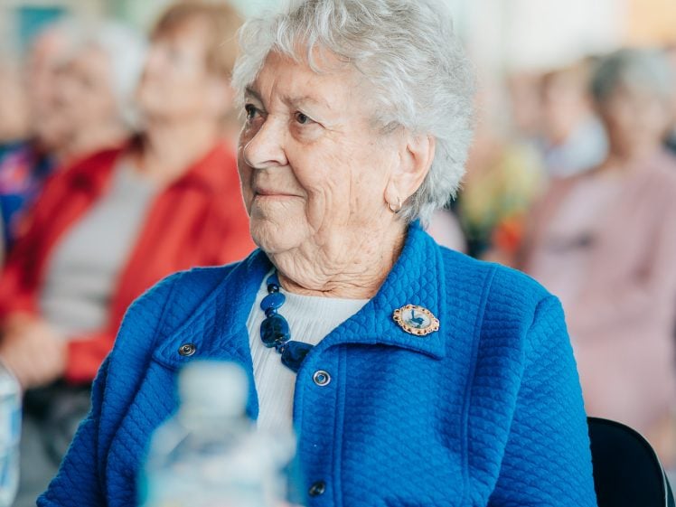 Image of An elderly woman with white hair and a blue jacket sits at a table, smiling. A water bottle and a banana are on the table in front of her. People are seated and blurred in the background.