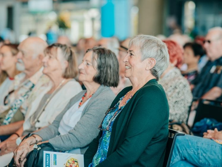 Image of A group of older adults sit closely together in an audience, attentively listening and smiling. One woman in the front holds a leaflet, and the setting appears to be a bright indoor event or seminar.