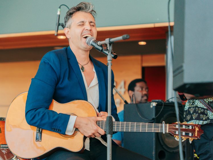 Image of A person in a blue blazer plays an acoustic guitar and sings into a microphone on stage, eyes closed with emotion. Musical equipment and another musician are visible in the background.