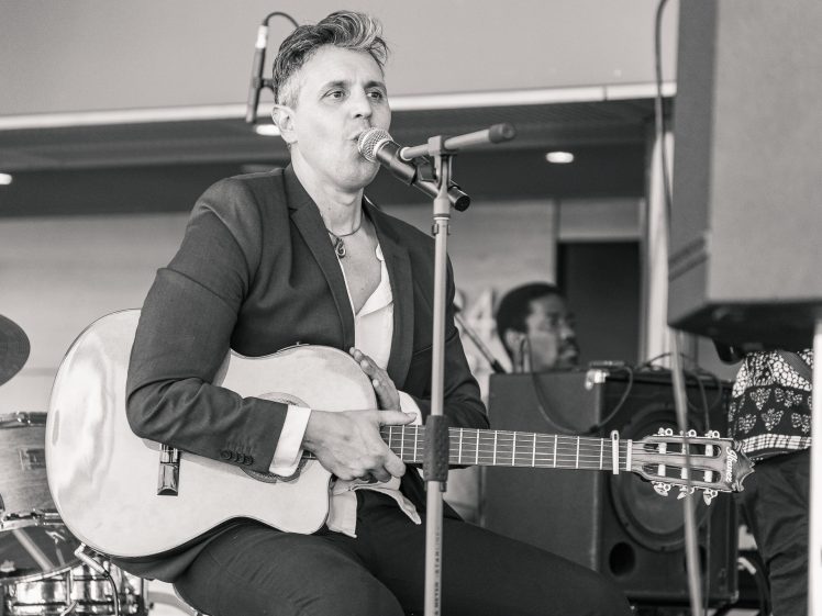 Image of A musician in a suit sits and sings into a microphone whilst holding an acoustic guitar. There are instruments and other performers in the background. The image is in black and white.
