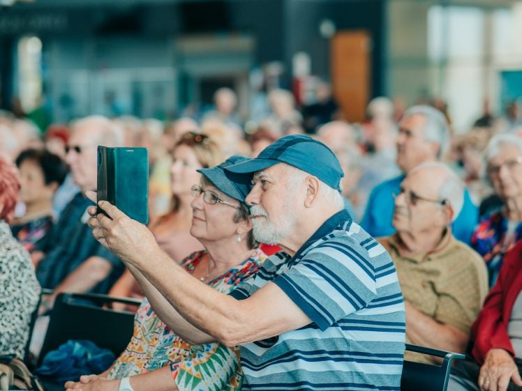 Image of An elderly man wearing a blue cap and striped shirt takes a photo with a tablet whilst sitting next to a woman in a floral top among a large audience of seated elderly people.