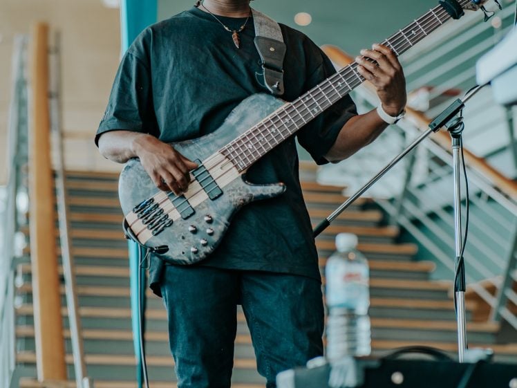 Image of A person in a black outfit plays a six-string bass guitar onstage in front of a staircase, with a microphone stand and water bottle nearby.