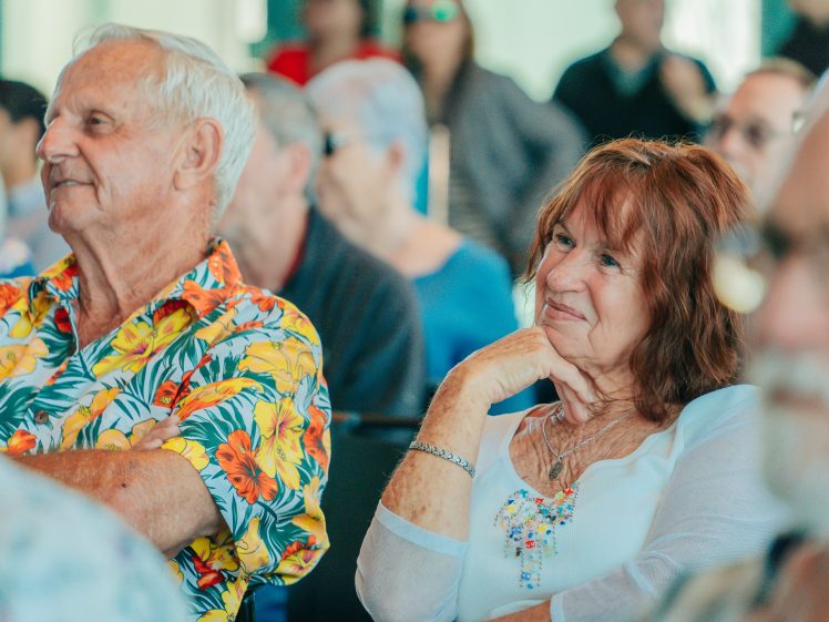 Image of An older man in a colourful floral shirt and an older woman in a white top sit smiling and listening attentively among a group of people at an indoor event.