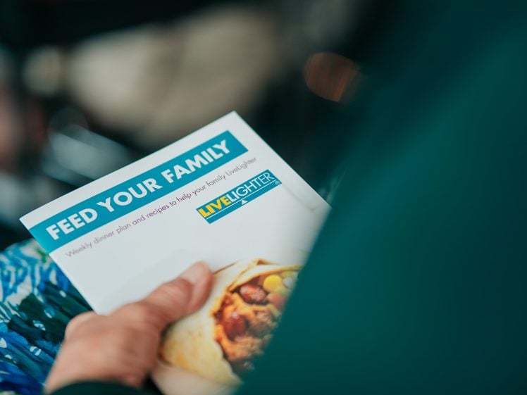 Image of A person holds a booklet titled FEED YOUR FAMILY with the LiveLighter logo, featuring a photo of a wrap sandwich on the cover. The booklet appears to include meal plans and recipes.