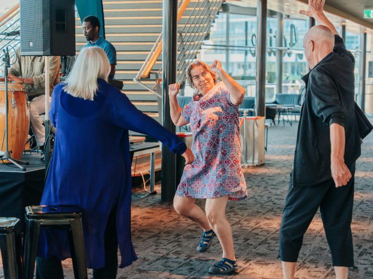 Image of Three people dance enthusiastically indoors near a musician playing the drums. The woman in the centre, wearing a patterned dress, smiles and poses, while the others dance beside her under bright natural light.