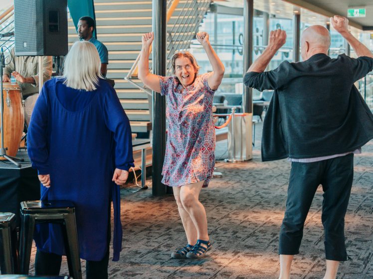 Image of Three adults dance energetically indoors; a smiling woman in a patterned dress stands between two others, raising her arms joyfully. The setting is modern with large windows and a staircase in the background.