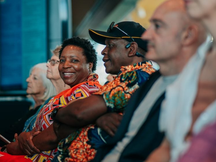 Image of A group of people sit closely together in a row, smiling and appearing engaged. The focus is on a woman in patterned clothing who looks directly at the camera whilst others face forward.