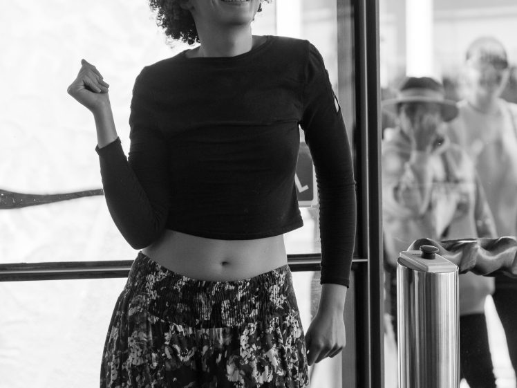 Image of A smiling person with curly hair, glasses, a long-sleeve black crop top, and patterned trousers stands by a glass door. Several people are visible outside through the glass. The image is in black and white.