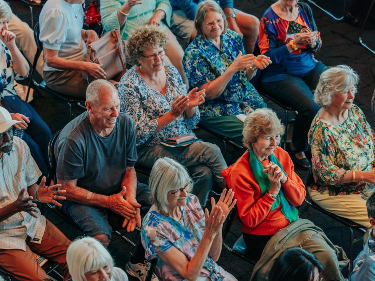 Image of A group of older adults sit in rows of chairs, smiling and clapping during an indoor event. They wear colourful clothing and appear to be engaged and enjoying the moment.