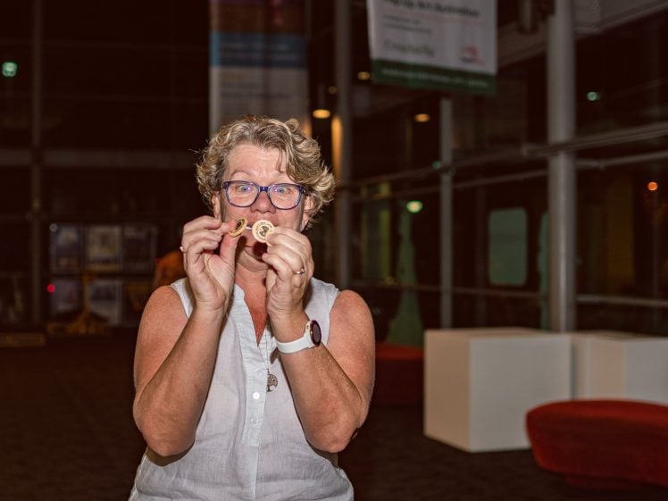 Image of A woman with short, curly hair and glasses, wearing a sleeveless white top and a watch, holds up two gold medals and smiles indoors, with modern furniture and glass walls in the background.