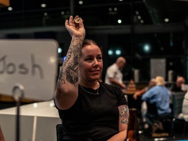 Image of A woman with tattoos on her arms holds up a poker chip while sitting at a table. A sign with the name Josh is visible, and there are people and tables in the background in a dimly lit room.
