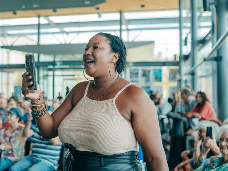 Image of A woman smiles and holds up her mobile to record or take a photo at an indoor event, surrounded by a seated crowd in a sunlit, modern venue.