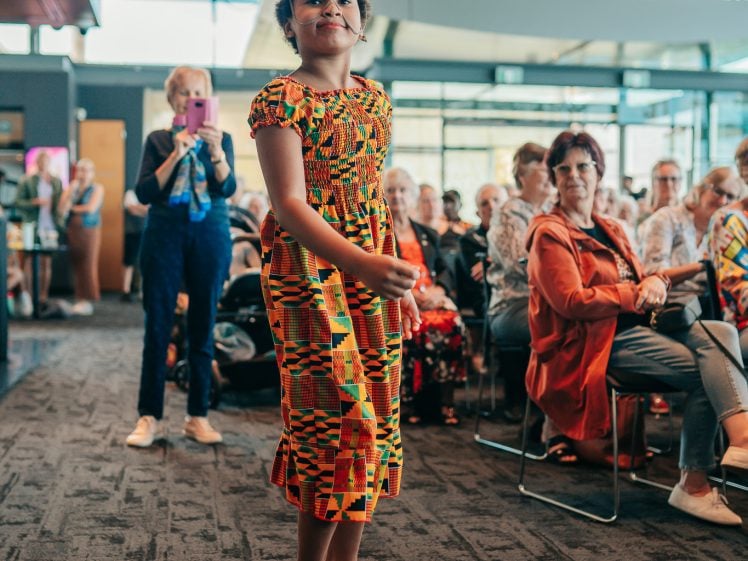 Image of A young girl in a colourful traditional dress and beaded headpiece dances confidently in front of a seated audience, while an older woman in the background takes a photo with her phone.