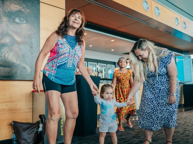 Image of Two women hold the hands of a small child as they stand indoors. Another child in a colourful dress walks in the background. The adults smile, and there is wall art and bright decor in the modern room.
