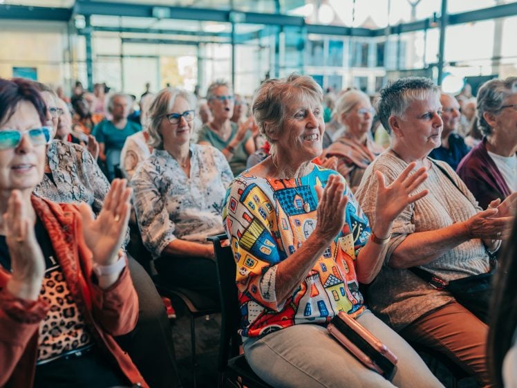 Image of A group of older adults sit closely together in rows, smiling and clapping during an indoor event. They appear engaged and cheerful, with natural daylight streaming through large windows in the background.