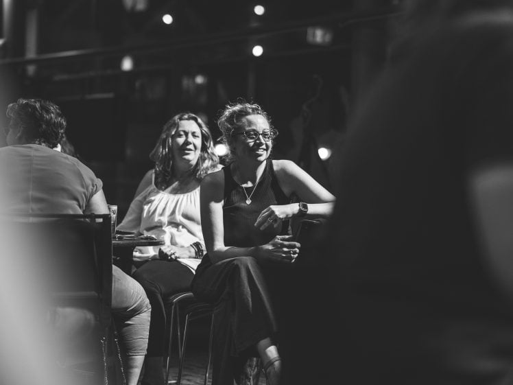 Image of Black and white photo of three women sitting at a table in a dimly lit setting, two are facing forwards and talking, one smiling, whilst a blurred figure passes by in the foreground.
