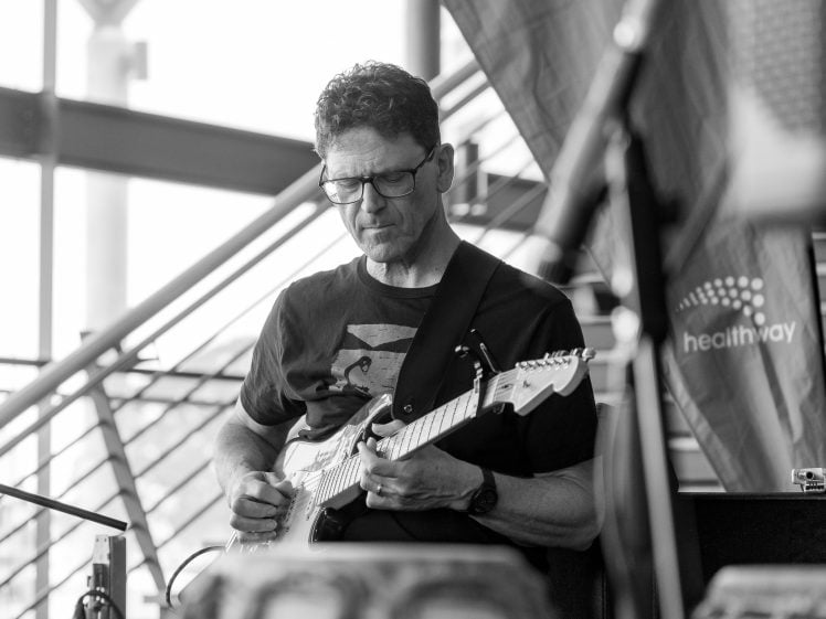 Image of A man wearing glasses and a T-shirt plays an electric guitar indoors. He appears focused on his playing, with stairs and a Healthway banner visible in the background. The photo is in black and white.