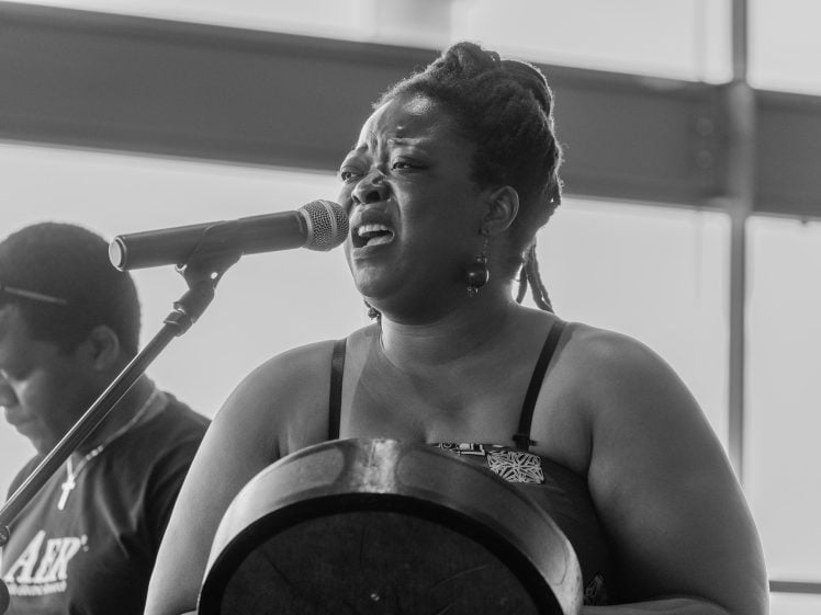 Image of A woman passionately sings into a microphone whilst playing a drum, with another person standing beside her in the background. The image is in black and white.