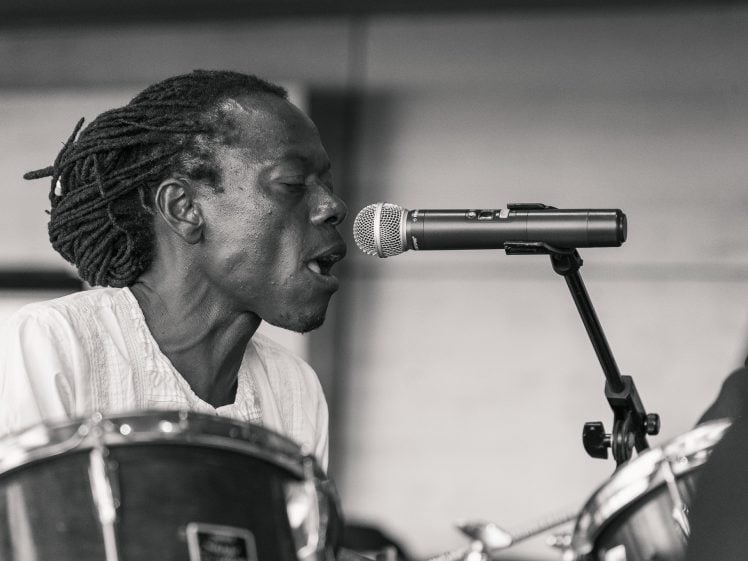 Image of A black-and-white photo of a drummer with dreadlocks, wearing a light shirt, passionately singing into a microphone whilst playing the drums. The background is softly blurred.