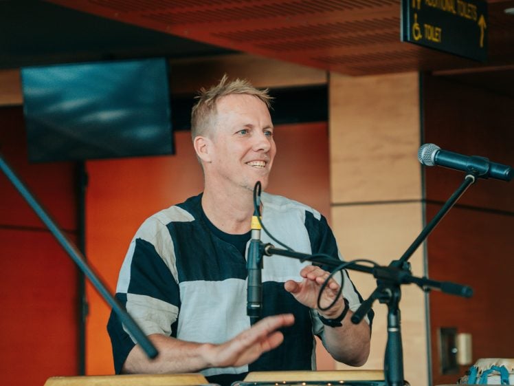 Image of A smiling man in a black and white shirt plays a drum and speaks or sings into a microphone, with another microphone and audio equipment visible in front of him indoors.