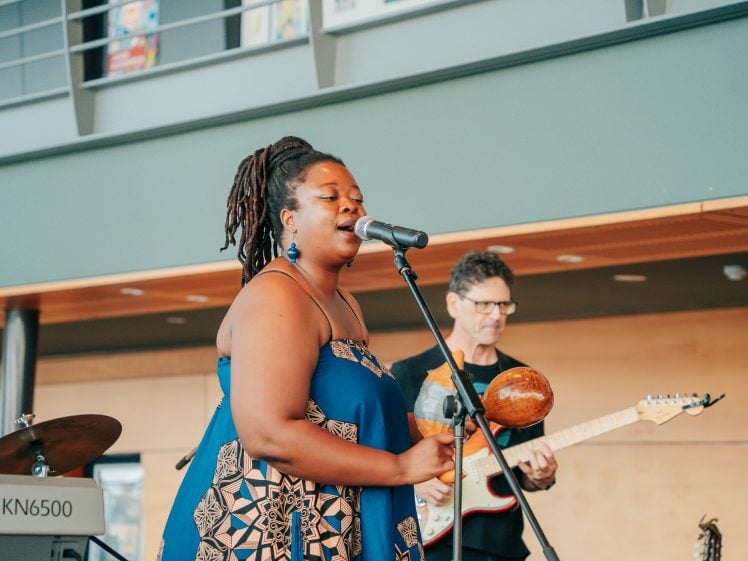 Image of A woman in a blue patterned dress sings into a microphone while holding a percussion instrument. A man in the background plays an electric guitar. They perform indoors near a keyboard and a balcony with posters above.