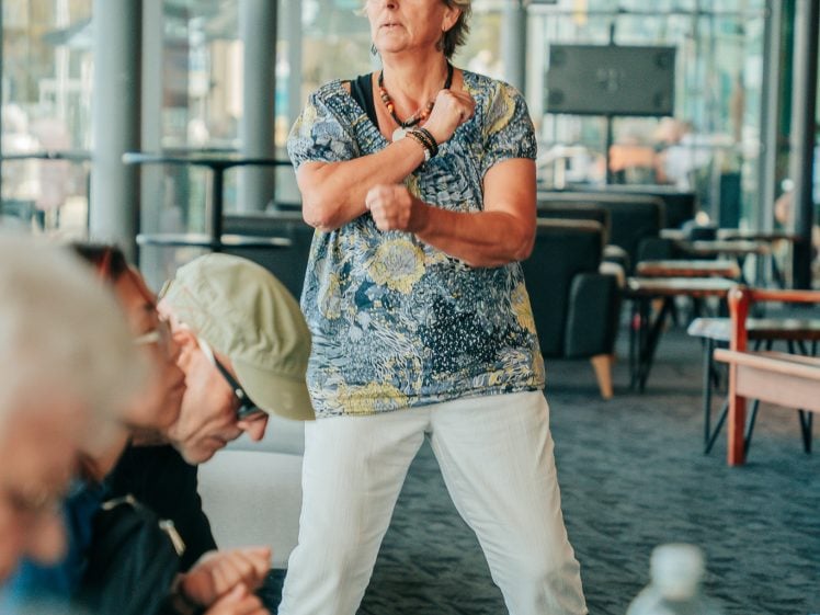 Image of An older woman stands and gestures with her arms in a modern, glass-walled room, whilst others sit at a table in the foreground. The scene suggests an engaging discussion or presentation.