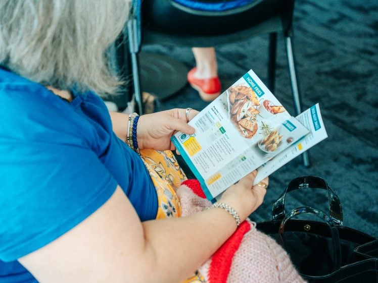 Image of A person with grey hair wearing a blue top and colourful skirt sits and reads a food menu, with a black handbag and red shoes visible nearby on a dark carpeted floor.