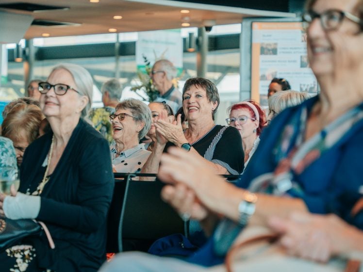 Image of A group of older adults sit together indoors, smiling and laughing. Some are clapping and appear to be enjoying a performance or presentation. The atmosphere is lively and joyful.