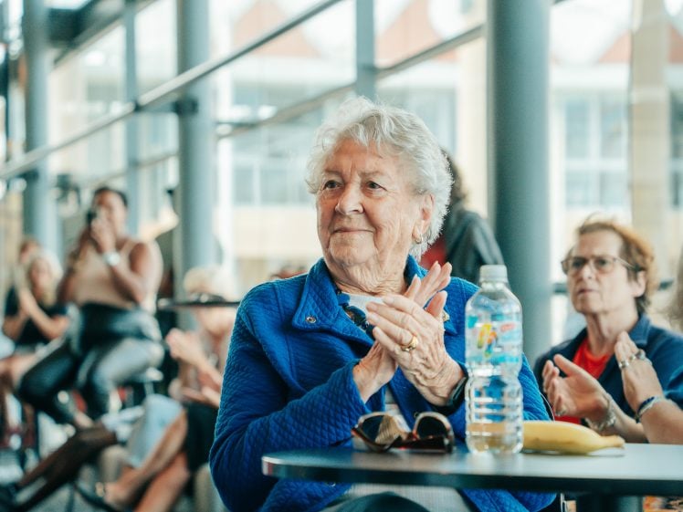 Image of An elderly woman in a blue jacket sits at a table, smiling and clapping. Sunglasses, a water bottle, and a banana are on the table. Other people are visible in the background in a bright, modern space.