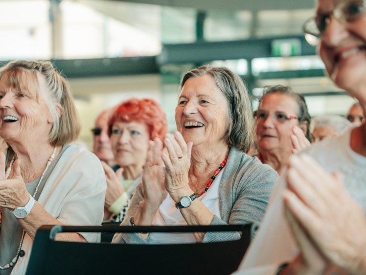Image of A group of older women sitting closely together indoors, smiling and clapping, appearing engaged and happy during an event or gathering.