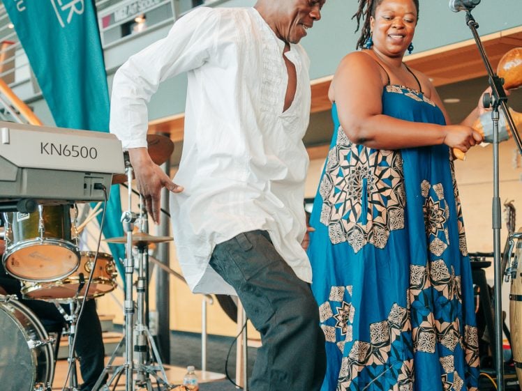 Image of A man in a white shirt and black trousers dances energetically next to a smiling woman in a blue patterned dress on a stage with musical instruments and a microphone.