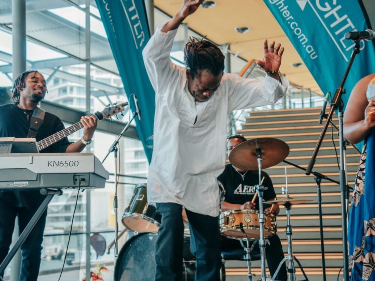 Image of A musician in a white tunic energetically dances on stage, surrounded by band members playing keyboard, drums, and guitar. Two blue LiveLighter banners and large windows are in the background.