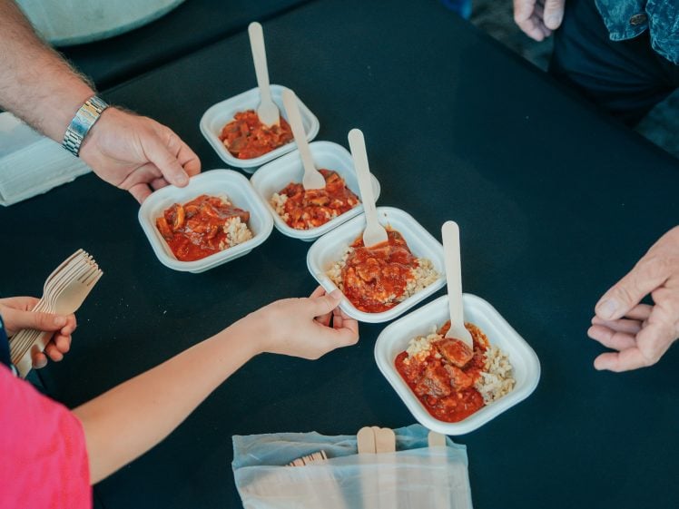 Image of Several white containers with rice and stew are being handed out on a dark table. Two people’s hands are reaching for the food, and wooden forks are visible in the foreground.