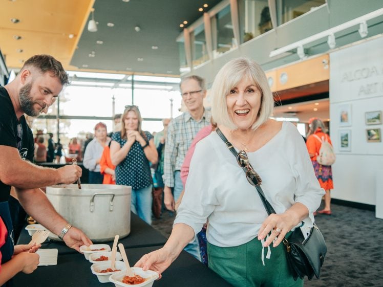 Image of A smiling woman holds a small dish of food at a counter whilst others wait in queue behind her at a lively indoor event with large windows and artwork on the walls.