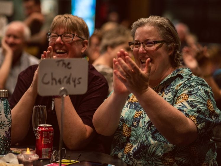 Image of Two people sit at a table labelled The Chardys, smiling and clapping enthusiastically in a lively, crowded indoor setting. Drinks, snacks, and a water bottle are visible on the table.